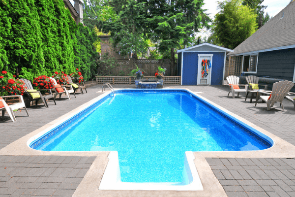 Backyard pool with cover off, blue water. Chairs on pool deck and greenery around.